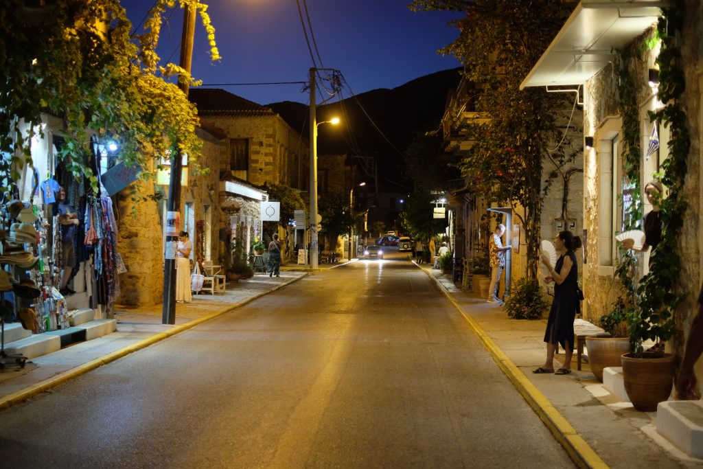 Narrow cobblestone street at dusk with warm streetlights illuminating stone buildings. People browse shops with vibrant textiles, creating a peaceful, inviting atmosphere.
