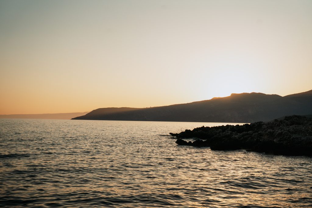 Sunset over calm sea with gentle waves, silhouetted rocky coastline, and softly glowing horizon creates a serene and peaceful atmosphere.