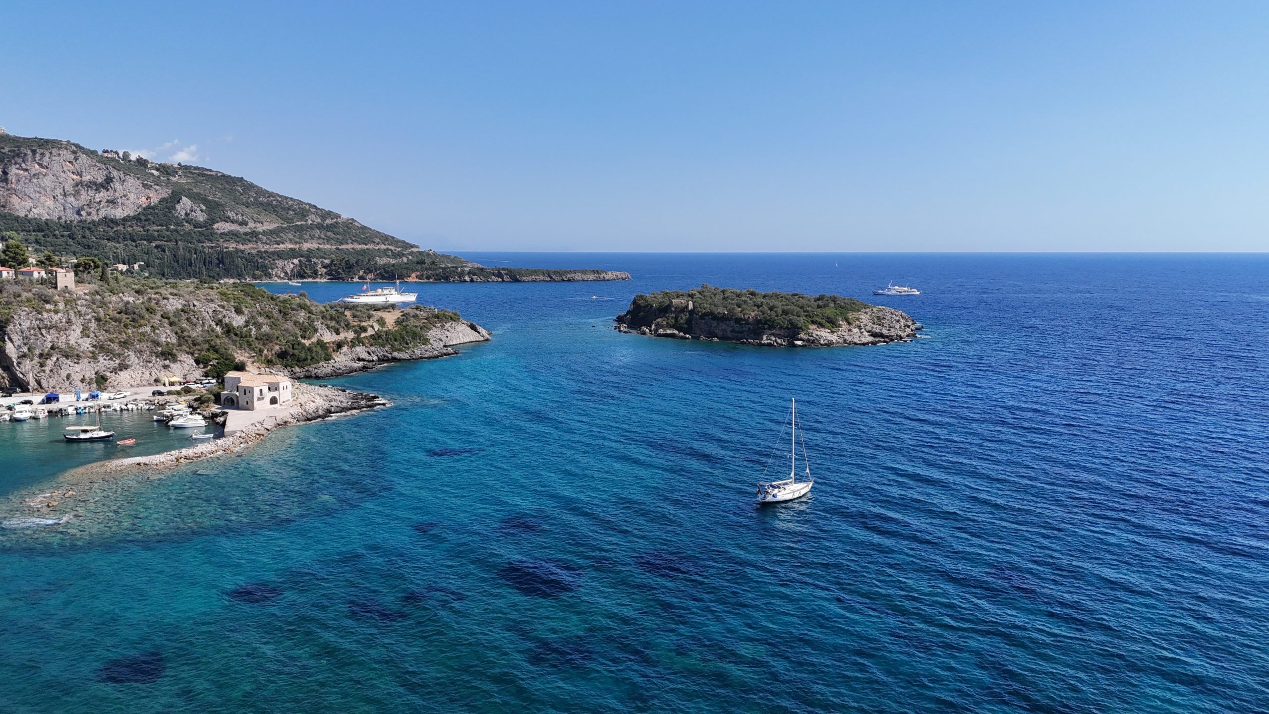 A serene coastal scene with a clear blue sea and a lone sailboat. Rocky green hills frame the bay under a bright blue sky, conveying tranquility and calm.