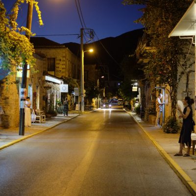 Narrow cobblestone street at dusk with warm streetlights illuminating stone buildings. People browse shops with vibrant textiles, creating a peaceful, inviting atmosphere.