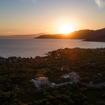 Aerial view of Kardamyli at sunset, showcasing the sea and distant houses bathed in warm, golden light.
